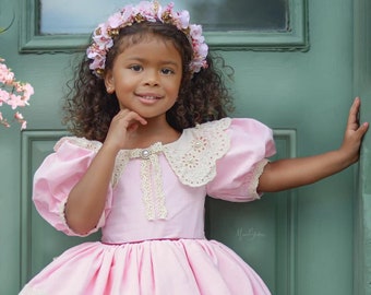May include: A young girl in a pink dress with a floral headband. The dress features a Peter Pan collar, puff sleeves, and a tiered skirt with lace trim. She wears white sandals and poses by a green door.