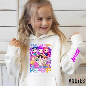 May include: A white hoodie featuring a colorful graphic of cartoon characters with the text "DEMON HUNTERS" and "KPOP". The right sleeve displays the word "NAME" in a bright pink font. The hoodie has a front pocket and a drawstring hood.
