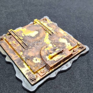May include: A weathered, square-shaped miniature with a rusted, metallic appearance. The surface is a mix of brown and gold, with visible bolts and a small, circular door. The edges are framed with a gray border, suggesting a model or diorama piece.