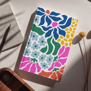 Puede incluir: Un cuaderno blanco con un dise&ntilde;o floral colorido. El dise&ntilde;o presenta flores y hojas rosas, azules, verdes, amarillas y naranjas.