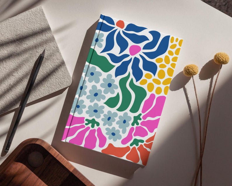 Puede incluir: Un cuaderno blanco con un dise&ntilde;o floral colorido. El dise&ntilde;o presenta flores y hojas rosas, azules, verdes, amarillas y naranjas.