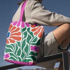 Puede incluir: Una bolsa de tela rosa con un dise&ntilde;o floral que presenta grandes flores naranjas y rojas, hojas verdes y flores rosas m&aacute;s peque&ntilde;as. La bolsa est&aacute; hecha de lona y tiene un asa rosa.