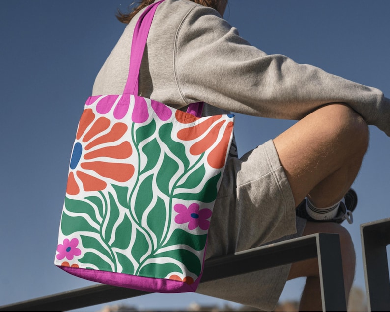 Puede incluir: Una bolsa de tela rosa con un dise&ntilde;o floral que presenta grandes flores naranjas y rojas, hojas verdes y flores rosas m&aacute;s peque&ntilde;as. La bolsa est&aacute; hecha de lona y tiene un asa rosa.