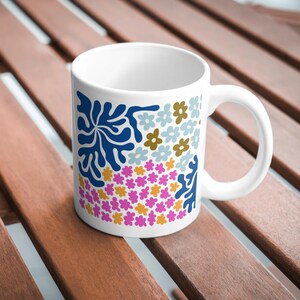 Puede incluir: Taza de cer&aacute;mica blanca con un dise&ntilde;o floral y abstracto colorido. El dise&ntilde;o presenta flores azules, rosas, amarillas y blancas, as&iacute; como una forma abstracta azul.