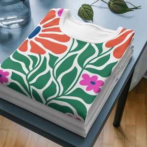 Puede incluir: Una camiseta blanca con un estampado floral verde, naranja, rosa y azul. La camiseta est&aacute; doblada y apilada sobre una mesa azul.