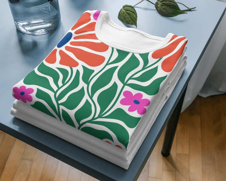 Puede incluir: Una camiseta blanca con un estampado floral verde, naranja, rosa y azul. La camiseta est&aacute; doblada y apilada sobre una mesa azul.