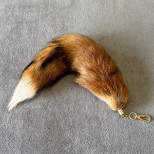 Può includere: Un portachiavi a forma di coda di volpe marrone e bianca con una catena dorata. La coda è soffice e realistica.