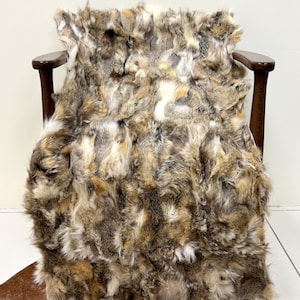 Può includere: Un plaid in finta pelliccia, nei toni del marrone, beige e crema, drappeggiato su una sedia di legno. La sedia ha una finitura in legno scuro. Il plaid è posto sopra un tappeto marrone. Il plaid in finta pelliccia è progettato per assomigliare alla pelliccia animale.