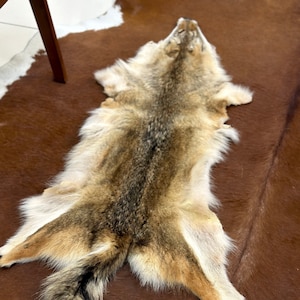 May include: A brown and grey wolf fur rug with a long tail. The fur is soft and fluffy, and the rug is laid out on a brown and white cowhide rug.