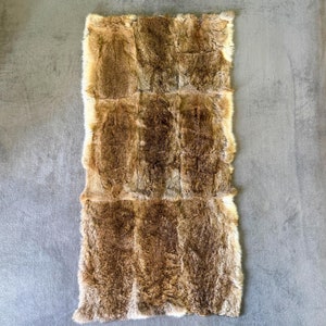 Puede incluir: Una alfombra rectangular de piel de conejo marrón. La piel es suave y esponjosa, y la alfombra está hecha de varias piezas de piel cosidas juntas.