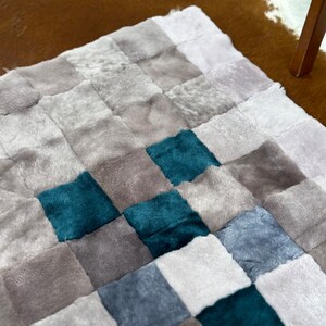 Op de afbeelding: Een pluche patchwork tapijt met een geruit patroon in grijstinten, blauw en teal.