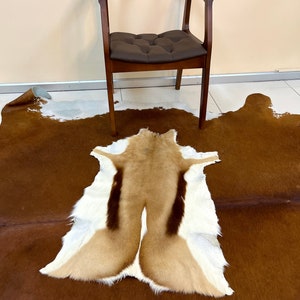 May include: A brown and white cowhide rug with a tan and white springbok hide rug on top. A dark brown wooden chair with a padded seat is in the background.