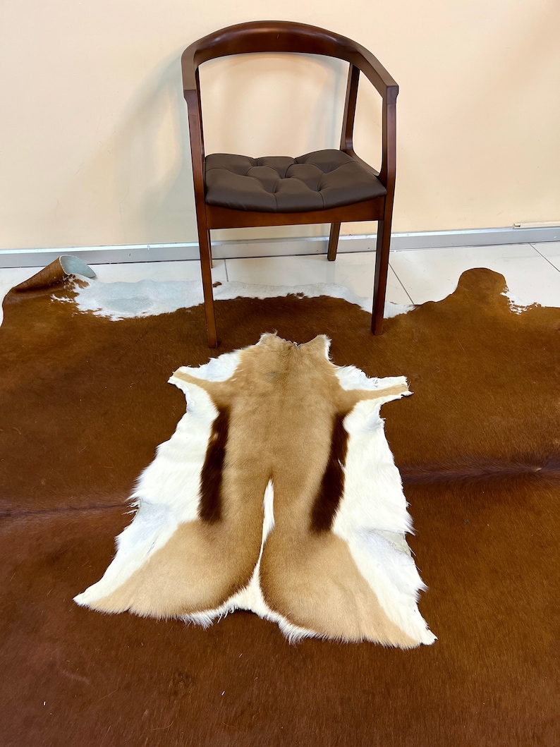 May include: A brown and white cowhide rug with a tan and white springbok hide rug on top. A dark brown wooden chair with a padded seat is in the background.