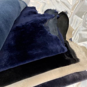 May include: A stack of different colored sheepskin hides. The top hide is a dark blue, the next is black, then a light beige, and the bottom is a dark gray.