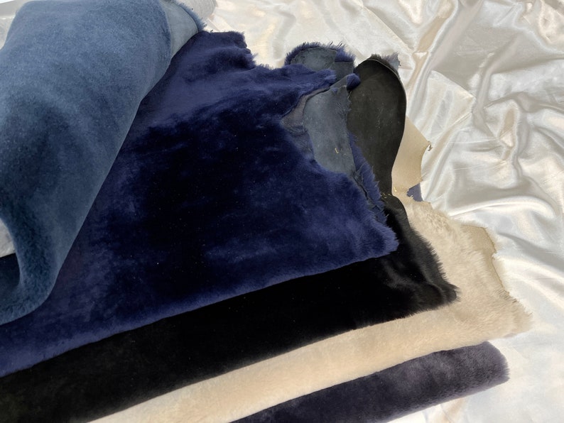 May include: A stack of different colored sheepskin hides. The top hide is a dark blue, the next is black, then a light beige, and the bottom is a dark gray.