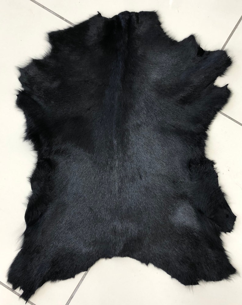 May include: A black cowhide rug with a natural, soft texture. The hide is laid flat on a white tiled surface.