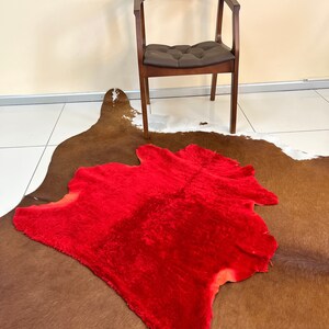 Peut inclure: Un tapis en fausse fourrure rouge sur un tapis en peau de vache brune et blanche. Une chaise en bois avec un si&egrave;ge en cuir brun est &agrave; l'arri&egrave;re-plan.