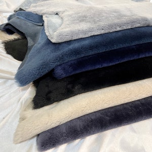 May include: A stack of different colored faux fur fabric swatches. The swatches are arranged in a pile, with the lightest color on top and the darkest color on the bottom. The colors are light blue, dark blue, black, cream, and dark gray.