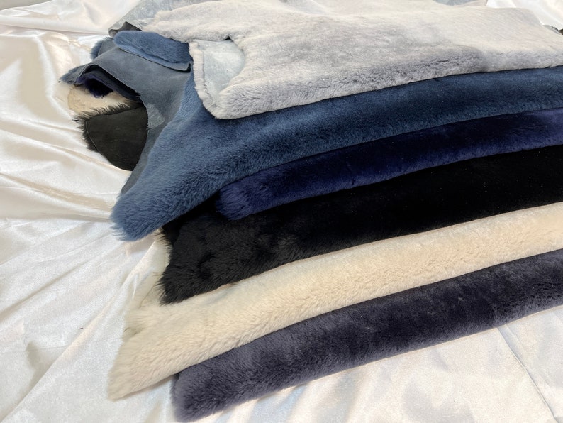 May include: A stack of different colored faux fur fabric swatches. The swatches are arranged in a pile, with the lightest color on top and the darkest color on the bottom. The colors are light blue, dark blue, black, cream, and dark gray.