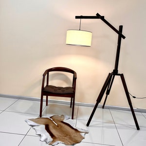 May include: A wooden tripod floor lamp with a white fabric lampshade. The lamp is next to a brown wooden chair with a brown cushion. A brown and white animal skin rug is on the floor.
