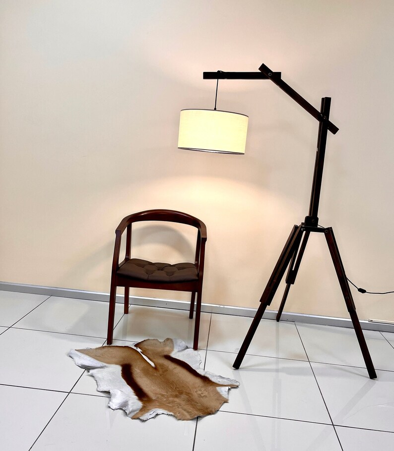 May include: A wooden tripod floor lamp with a white fabric lampshade. The lamp is next to a brown wooden chair with a brown cushion. A brown and white animal skin rug is on the floor.