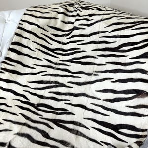 May include: A textile with a bold zebra stripe pattern. The fabric displays black stripes against a creamy white backdrop. The textile is draped over a white surface, highlighting its texture and design. The pattern is reminiscent of a zebra's coat.