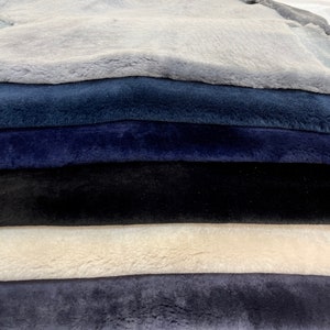 May include: A stack of six sheepskin rugs in various shades of blue and gray. The top rug is light gray, the second is a darker shade of blue, the third is a deep navy blue, the fourth is black, the fifth is a light beige, and the bottom rug is a medium shade of blue.