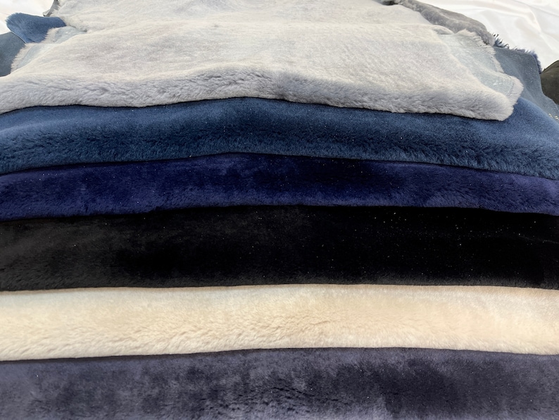 May include: A stack of six sheepskin rugs in various shades of blue and gray. The top rug is light gray, the second is a darker shade of blue, the third is a deep navy blue, the fourth is black, the fifth is a light beige, and the bottom rug is a medium shade of blue.