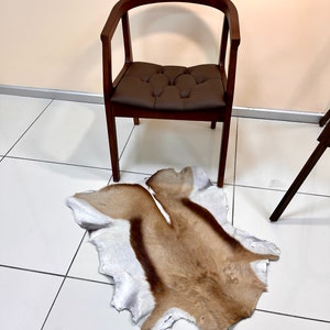 Op de afbeelding: Een bruine leren stoel met een gestoffeerde zitting staat op een witte tegelvloer met een bruin en wit dierenvachtkleed ervoor.