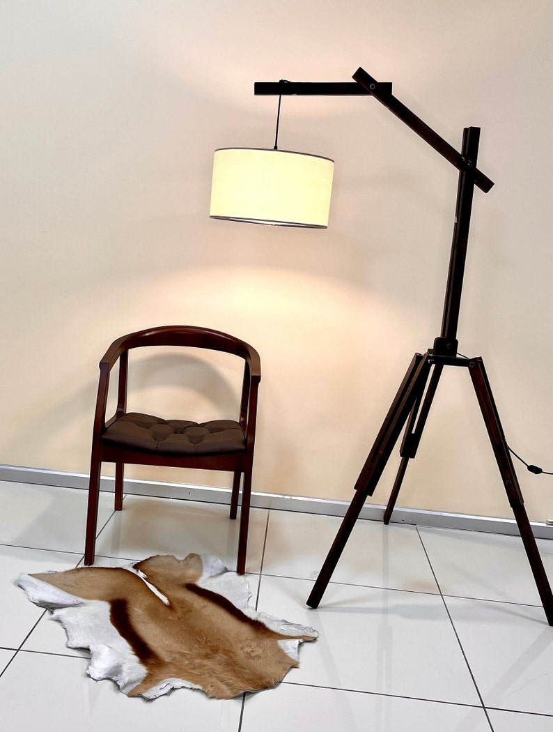Op de afbeelding: Een houten vloerlamp met een driepootbasis en een witte stoffen kap. De lamp staat naast een bruine stoel en een bruin en wit dierenhuidkleed.