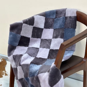 Op de afbeelding: Een geruite deken in grijstinten, blauw en wit. De deken is gemaakt van zacht, pluche bont en is over een houten stoel gelegd.