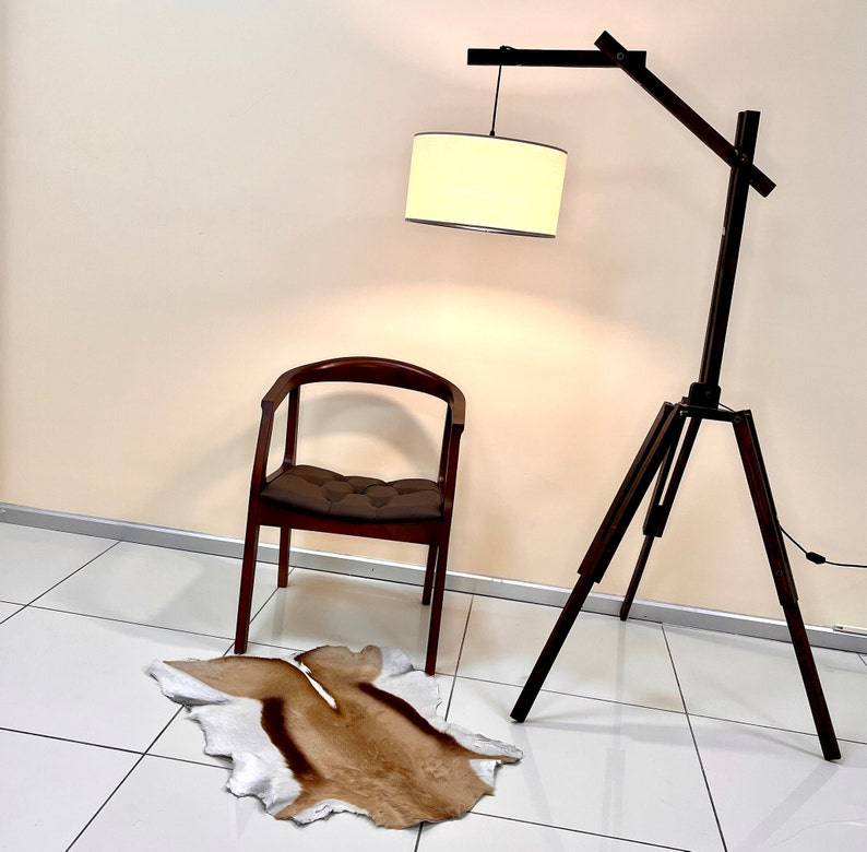 Op de afbeelding: Een bruine houten staande lamp met een witte lampenkap van stof. De lamp staat naast een bruine houten stoel met een bruin kussen. Een bruin en wit dierenvachtkleed ligt op de vloer.