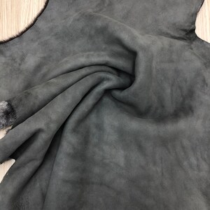 以下が含まれることがあります： 柔らかくふわふわの質感の濃いグレーの羊の皮。皮は折り畳まれ、明るい木製の表面にかけられています。