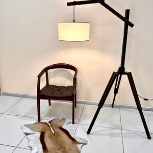 May include: A wooden floor lamp with a tripod base and a long, adjustable arm. The lamp has a white fabric shade and is lit. A brown leather chair and a brown and white animal skin rug are in the foreground.