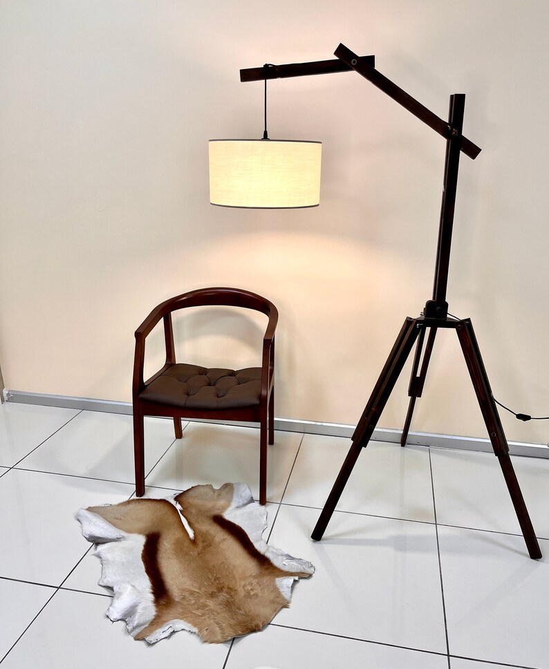 May include: A wooden floor lamp with a tripod base and a long, adjustable arm. The lamp has a white fabric shade and is lit. A brown leather chair and a brown and white animal skin rug are in the foreground.