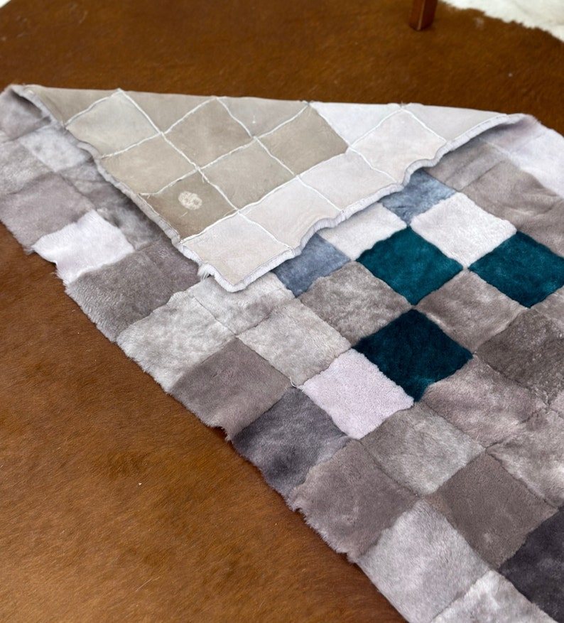 Może przedstawiać: Dywan patchwork wykonany z kwadrat&oacute;w z imitacji futra w r&oacute;żnych odcieniach szarości i turkusu. Dywan jest złożony na p&oacute;ł, odsłaniając pod spodem beżowy wz&oacute;r w kratkę.
