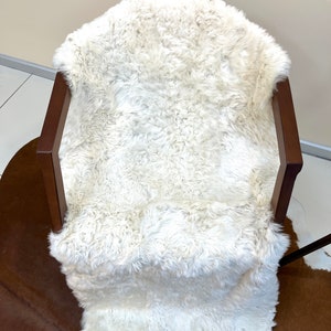 Puede incluir: Una alfombra de piel de oveja blanca drapeada sobre una silla de madera. La alfombra es suave y esponjosa, y la silla es de madera oscura.