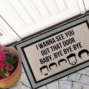 May include: A grey doormat with black text that reads "I wanna see you out that door baby, bye bye bye" with a row of black silhouettes of men's heads with different hairstyles.