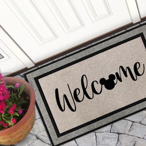May include: A welcome mat with a black and white border and a beige background. The text "Welcome" is written in black cursive script with a black Mickey Mouse silhouette in the middle of the word.