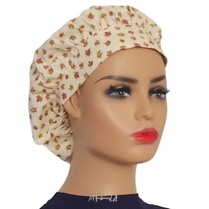May include: A beige fabric bouffant cap with a repeating pattern of small red and orange leaves. The cap is gathered at the back and has a rounded shape. The cap is worn by a mannequin.
