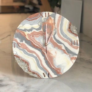 May include: A round clock with a white, gray, and pink marbled design. The clock face has Roman numerals and a gold-colored hour hand. The clock is decorated with small, clear crystals.