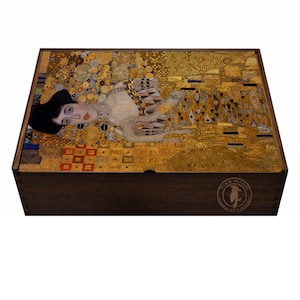Puede incluir: Caja de rompecabezas de madera con la pintura "Dama con abanico" de Gustav Klimt. La obra muestra a una mujer con un vestido dorado sobre un fondo dorado. La caja es de color marrón oscuro con un logotipo en la esquina inferior derecha.