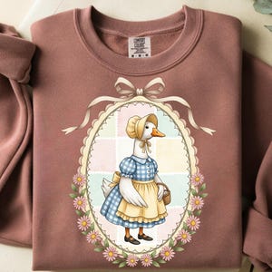 Peut inclure: Sweat-shirt Comfort Colors rose poussiéreux avec une illustration fantaisiste d'une oie vêtue d'une robe vichy bleue et d'un tablier jaune. L'oie est dans un cadre ovale avec une bordure florale et un nœud. Le sweat-shirt a un col rond.