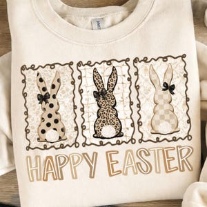 Könnte beinhalten: Cremefarbenes Sweatshirt mit drei Hasenmotiven in dekorativen Rahmen. Die Hasen haben Punkte-, Leopardenmuster- und Karomuster, jeweils mit einer schwarzen Schleife. Die Worte "HAPPY EASTER" sind in einer braunen, stilisierten Schrift gedruckt.