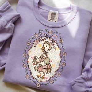 Puede incluir: Una sudadera lila Comfort Colors con una ilustración vintage de un ganso con gorro y vestido, sosteniendo un bolso y un huevo. El gráfico está enmarcado por un óvalo floral con un lazo rosa.