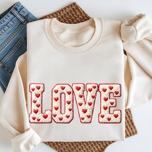 May include: Cream-colored sweatshirt with the word "LOVE" embroidered in a textured, off-white fabric. Each letter is adorned with small red hearts. The sweatshirt is folded, with a pair of blue jeans visible in the background.