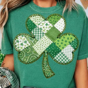 May include: Green t-shirt featuring a patchwork shamrock design. The shamrock is made of various green patterned fabrics, including polka dots, floral, and plaid. The edges of the shamrock are outlined with a decorative stitch.