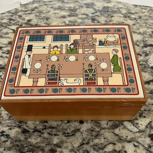 May include: A rectangular wooden box with a decorative tile lid featuring a colorful illustration of a family seated at a dining table. The border of the tile displays a repeating pattern of plates, forks, and knives in blue and red tones.