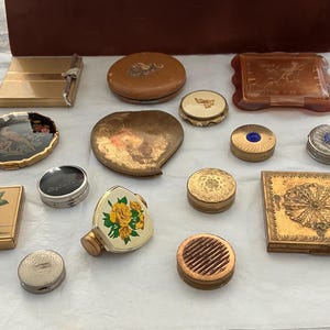 May include: A collection of vintage compacts and cosmetic containers in various shapes and sizes. The items are made of metal, glass, and other materials, featuring decorative designs and patterns. Colors include gold, brown, and silver.