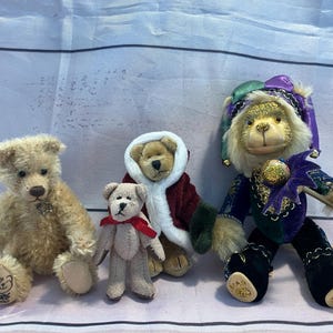 May include: Four plush teddy bears of varying sizes. One is light brown with a star pendant, another is small and tan with a red bow, and a third is wearing a red and white coat. The fourth bear is dressed in a jester costume with a purple and green hat.